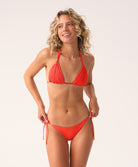 A woman wearing a red macrame triangle top bikini and tie side bottoms standing against a white wall.