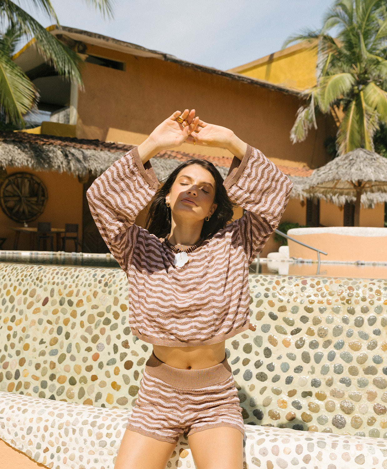 Woman in a brown and beige striped outfit posing outdoors with palm trees and a building in the background