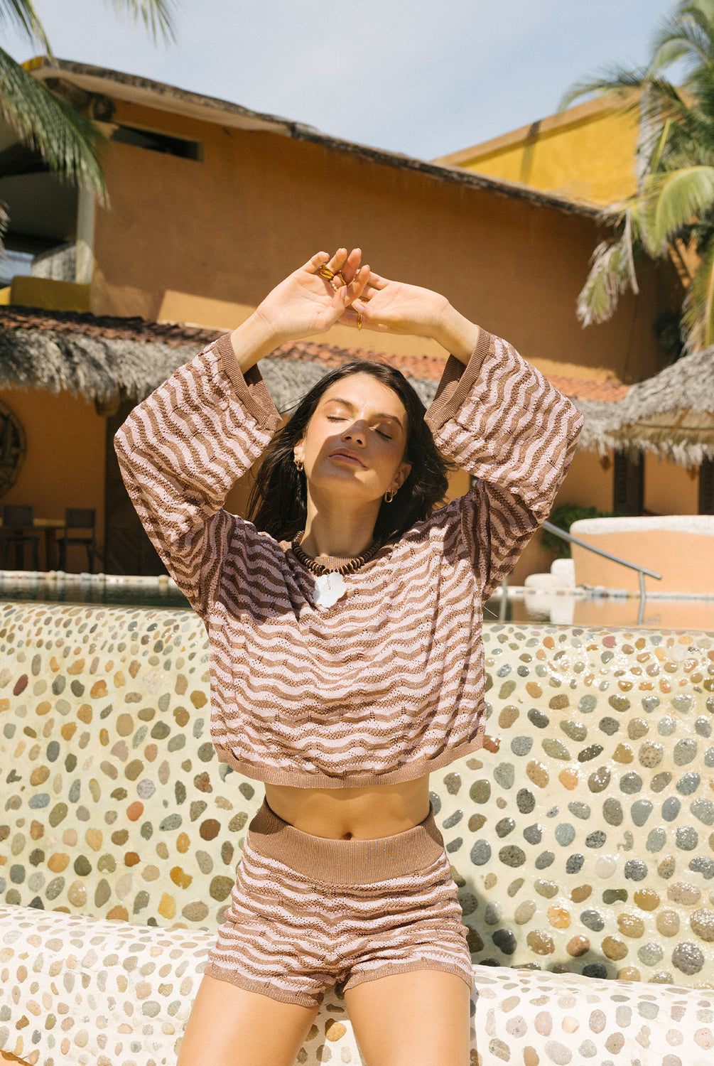 Woman in a brown and beige striped outfit posing outdoors with palm trees and a building in the background