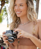 A woman wearing a multi colored necklace with a white pearl standing outside holding a camera.