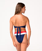 A woman wearing a red, white, and navy bandeau bikini facing towards a white wall.