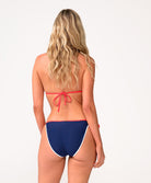 A woman wearing a navy triangle top bikini facing towards a white wall.