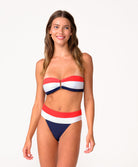 A woman wearing a red, white, and navy bandeau bikini standing against a white wall.