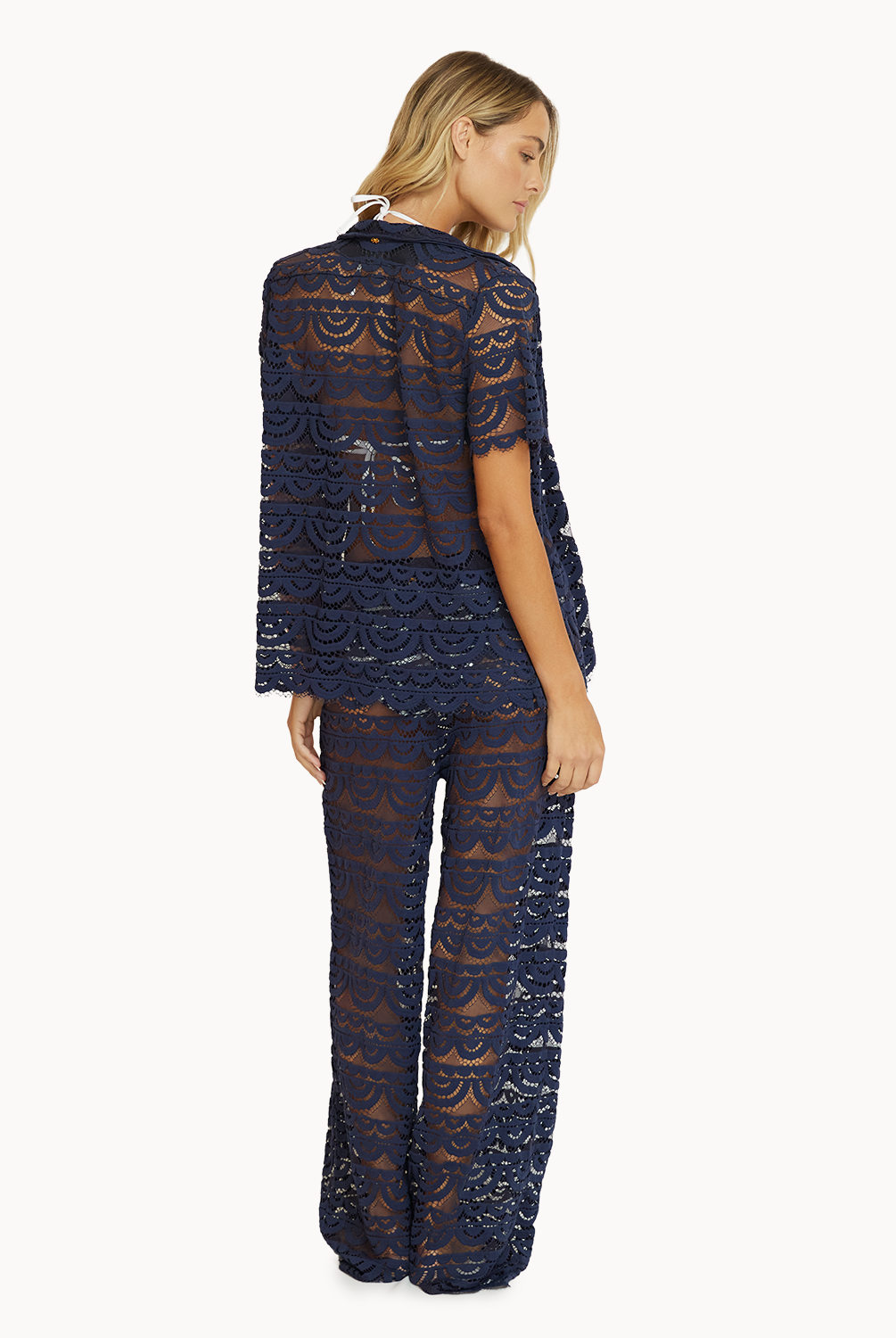 A woman wearing a navy lace short sleeve shirt and matching lace pants facing towards a white wall.