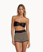 A woman wearing a black bikini and striped shorts standing against a white wall.