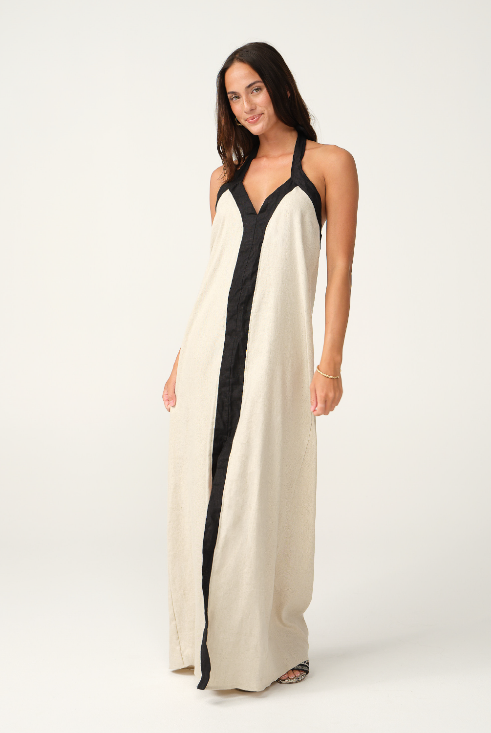 A woman modeling a long cream linen maxi dress with black trim and a black front placket, photographed against a seamless white studio background.