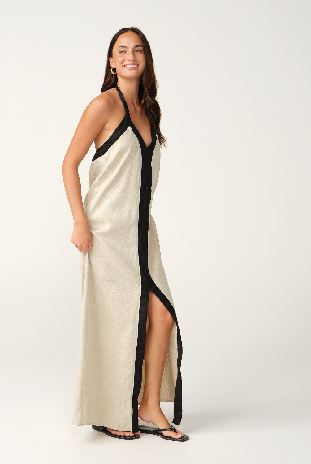 A woman modeling a long cream linen maxi dress with black trim and a black front placket, photographed against a seamless white studio background.