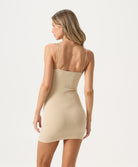 A woman wearing a nude slip dress facing towards a white wall.