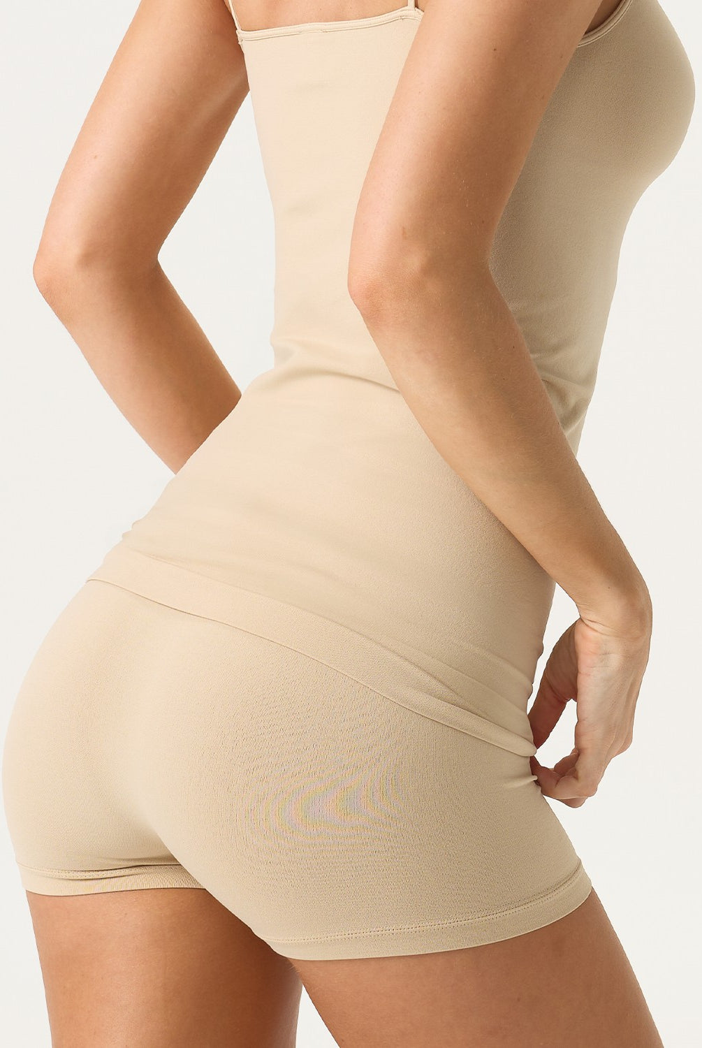 A woman wearing a nude tank top and matching shorts standing against a white wall.