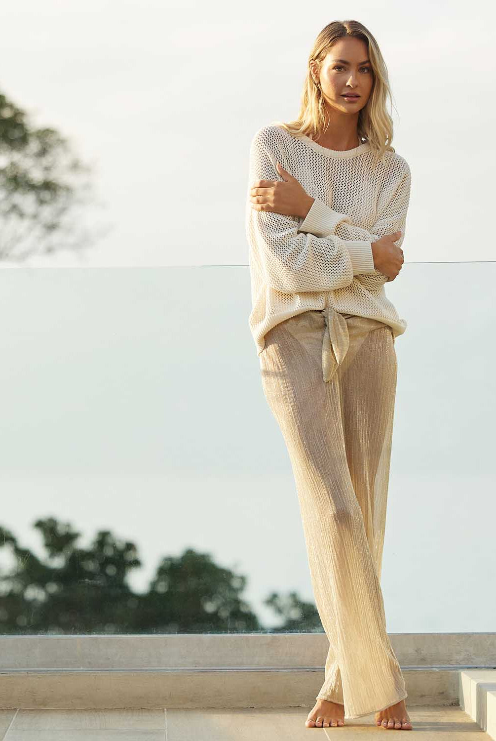 A blonde woman wearing an ivory knit sweater and long gold pants standing on a patio outside.