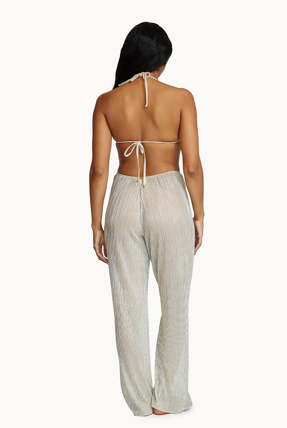 A woman wearing a bikini top and long gold pants facing towards a white wall.