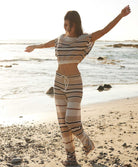 A woman wearing a matching striped crop top and long pants walking alongside the beach.