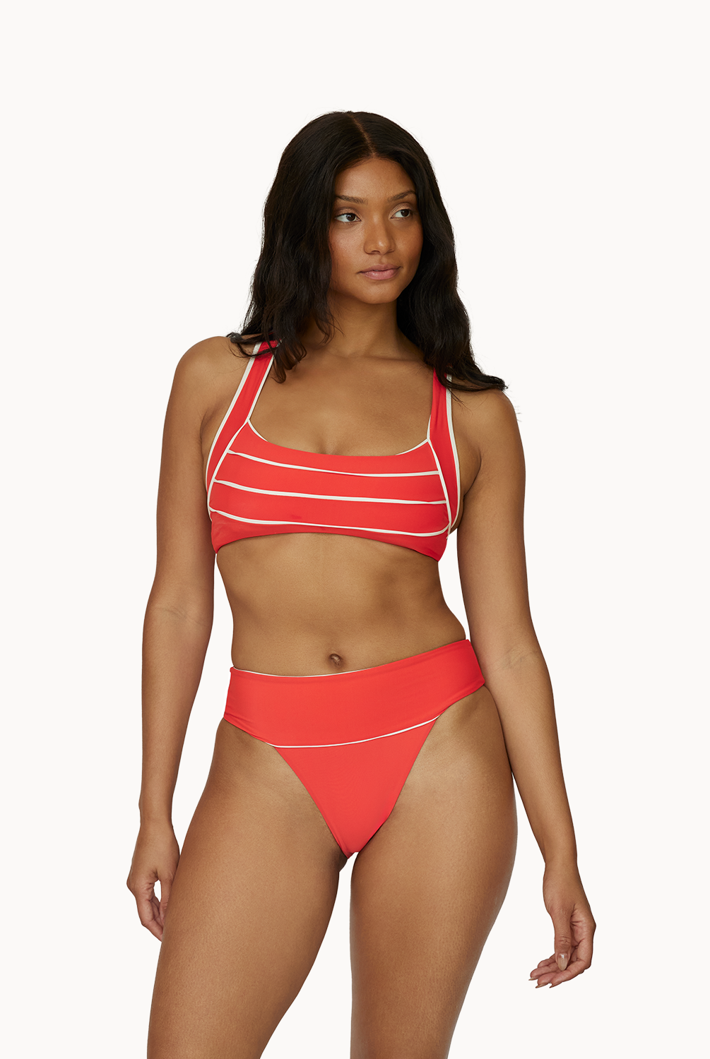 A woman wearing a red bikini with white piped detailing standing in front of a white wall.