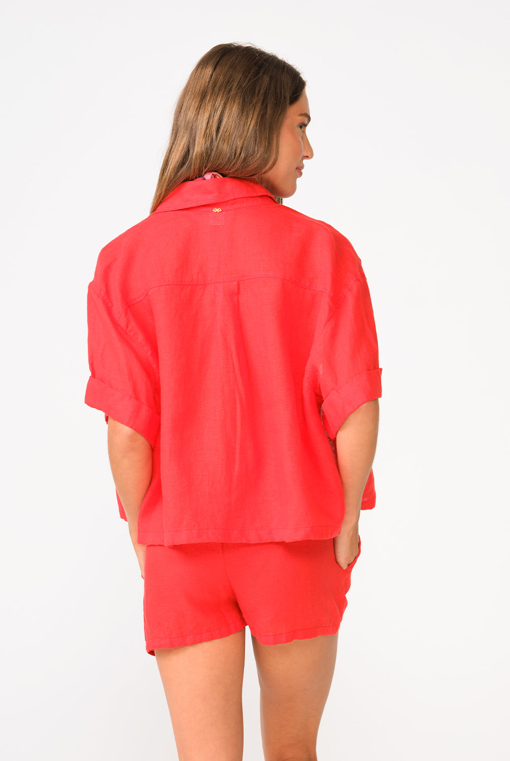 A woman wearing a red short sleeve linen top and matching linen shorts facing towards a white wall.