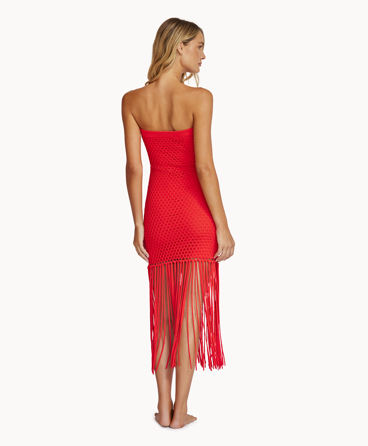 A woman wearing a red strapless mesh dress with fringe facing towards a white wall.