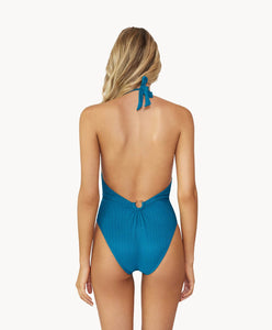 Blonde woman wearing a textured turquoise halter one-piece swimsuit with gold details facing backwards towards a white wall.