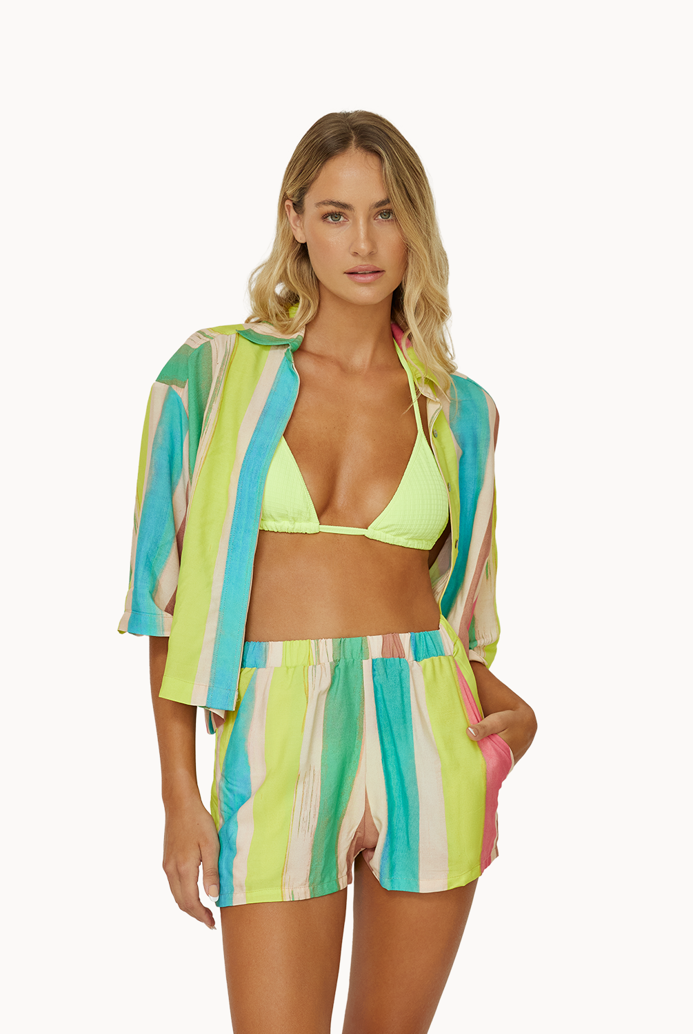 A woman wearing a green bikini top and a color block shirt and short set standing against a white wall.