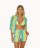 A woman wearing a green bikini top and a color block shirt and short set standing against a white wall.