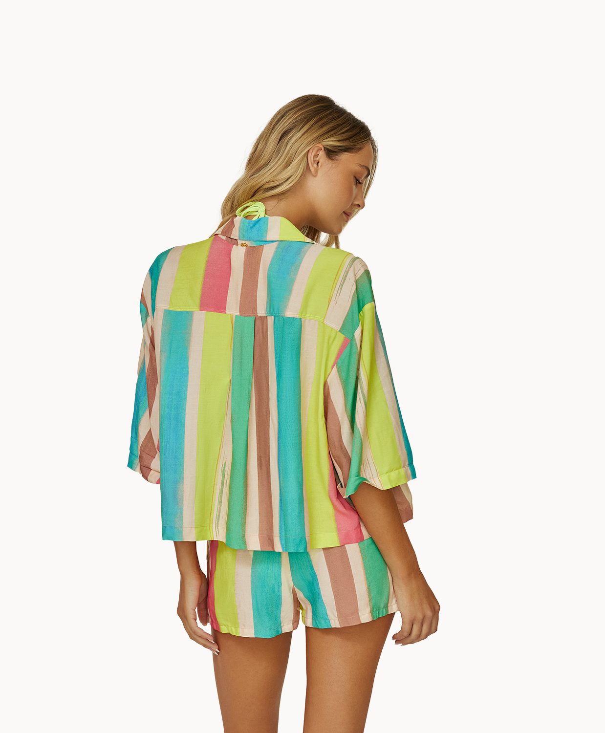A woman wearing a green bikini top and a color block shirt and short set facing towards a white wall.