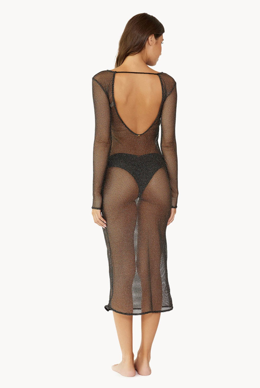 Brunette woman wearing a black sheer knee length coverup dress with sleeves over a black bikini facing backwards towards white wall.