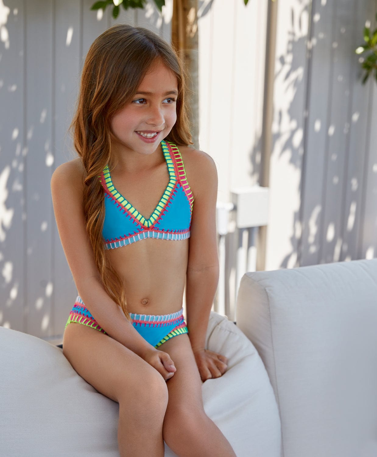 A brunette girl wearing a turquoise bikini with colorful embroidery sitting on a couch outside.