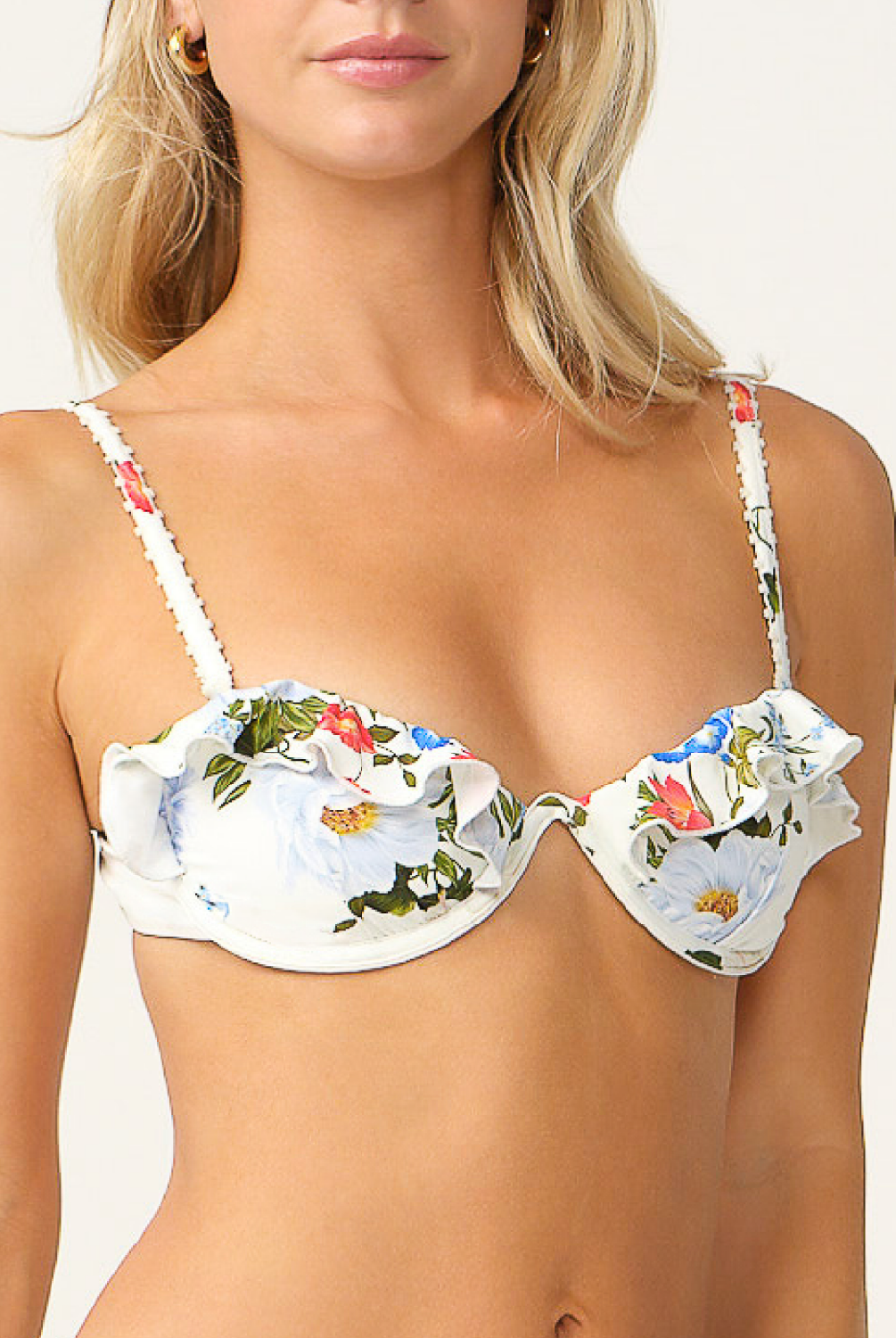 A close-up of a model wearing a white ruffled bikini top with a colorful floral print, featuring blue, pink, and green flowers with yellow centers.