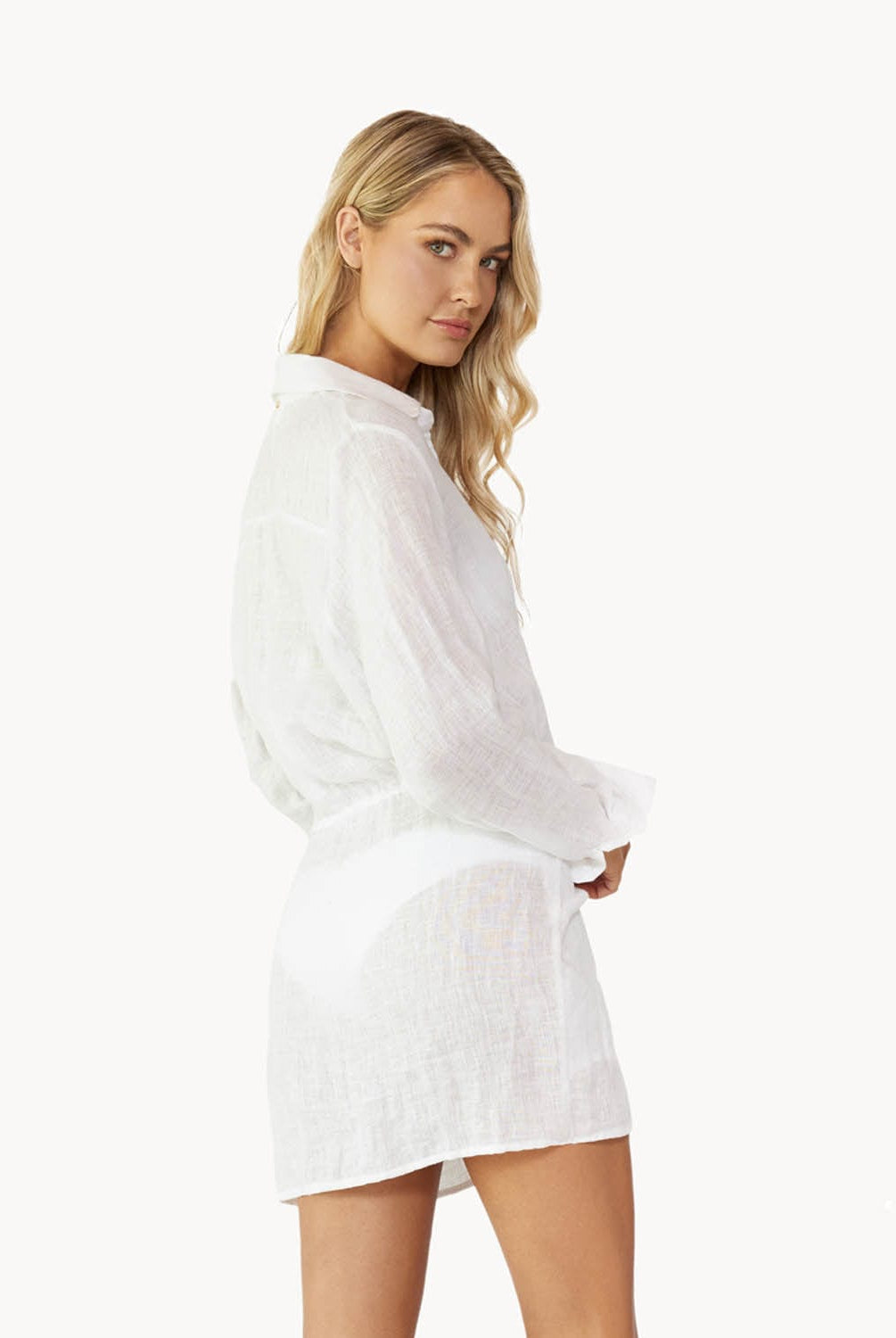 Blonde woman wearing a lightweight white linen button coverup with front-tie detail facing backwards towards white wall.