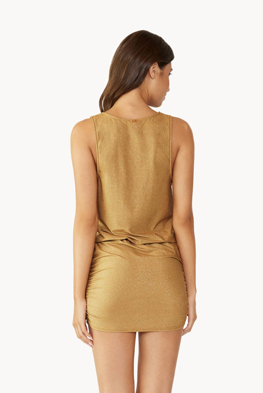 Brunette woman wearing a gold short coverup dress with front tie detail facing backwards towards white wall.