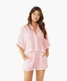 Brunette woman wearing a pink linen button coverup shirt and shorts stands in front of white wall.