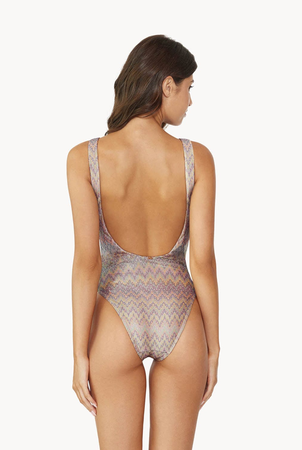 Brunette woman wearing a multi-colored print one piece swimsuit facing backwards towards a white wall.