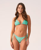 A woman wearing a teal triangle top bikini and matching tie side bottoms standing in front of a white wall.