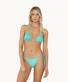 A blonde woman wearing a turquoise bikini stands in front of a white wall.
