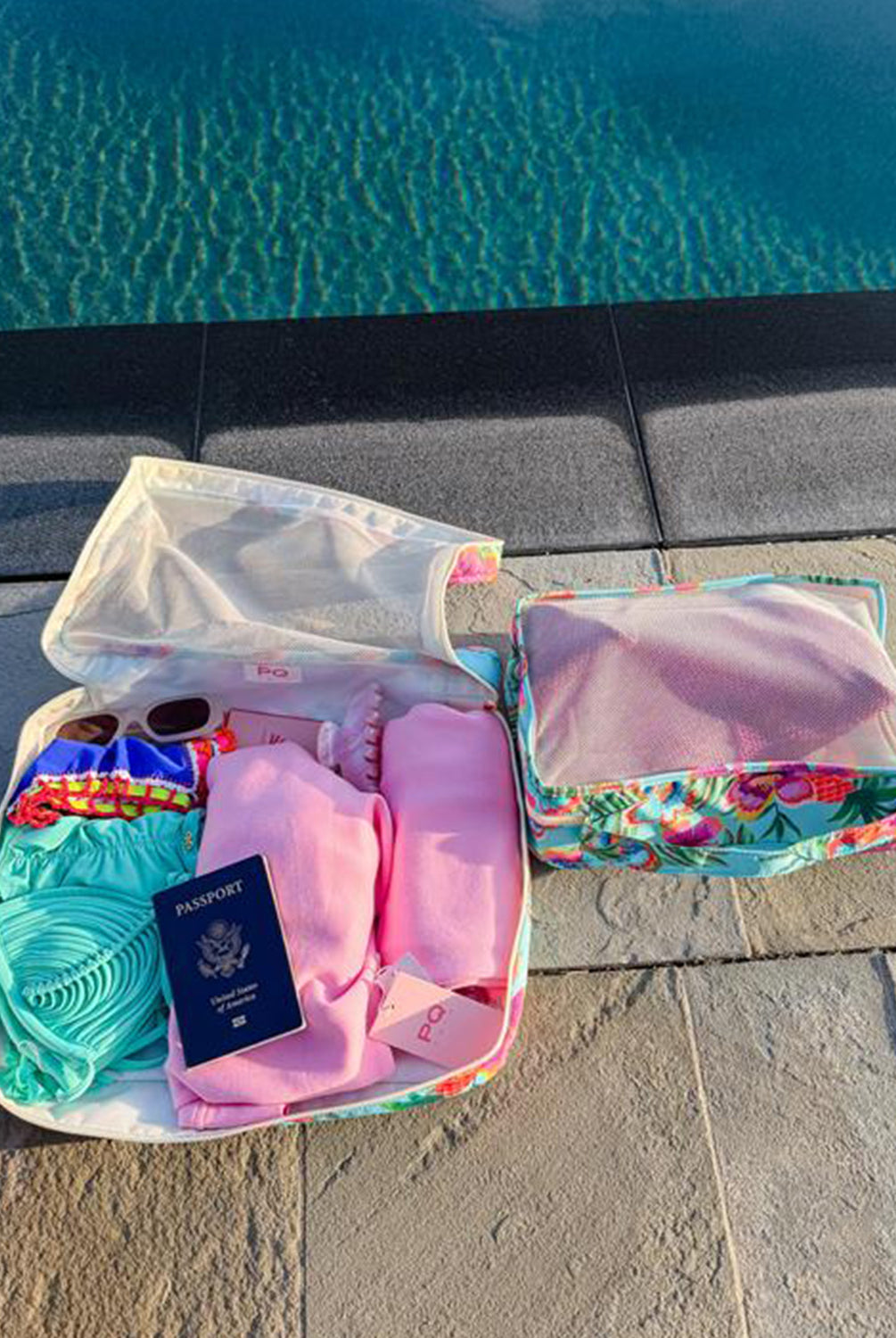 Open packing cube with travel items including a passport, sunglasses, and clothes by a pool.