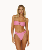 A blonde woman wearing a pink bikini standing against a white wall.