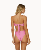 A blonde woman wearing a pink bikini facing towards a white wall.