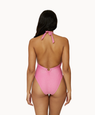 A woman wearing a pink one piece swimsuit facing towards a white wall.