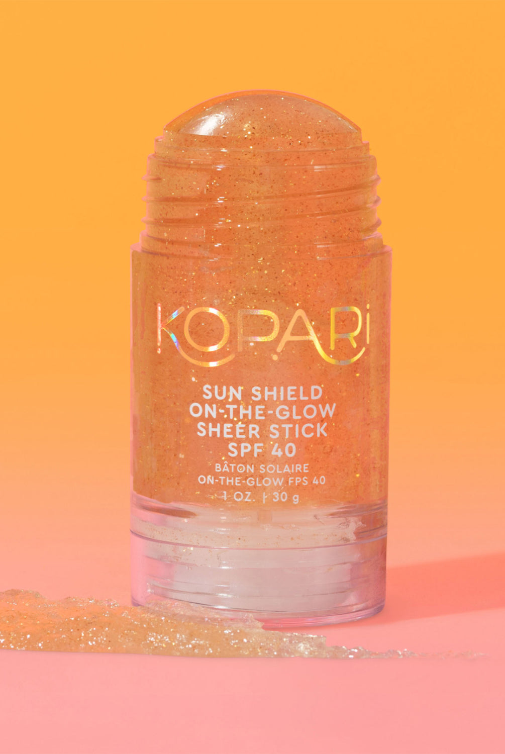 The Kopari Sun Shield On-the-Glow Sheer Stick SPF 40 standing upright with a swatch of product on an orange-pink background.