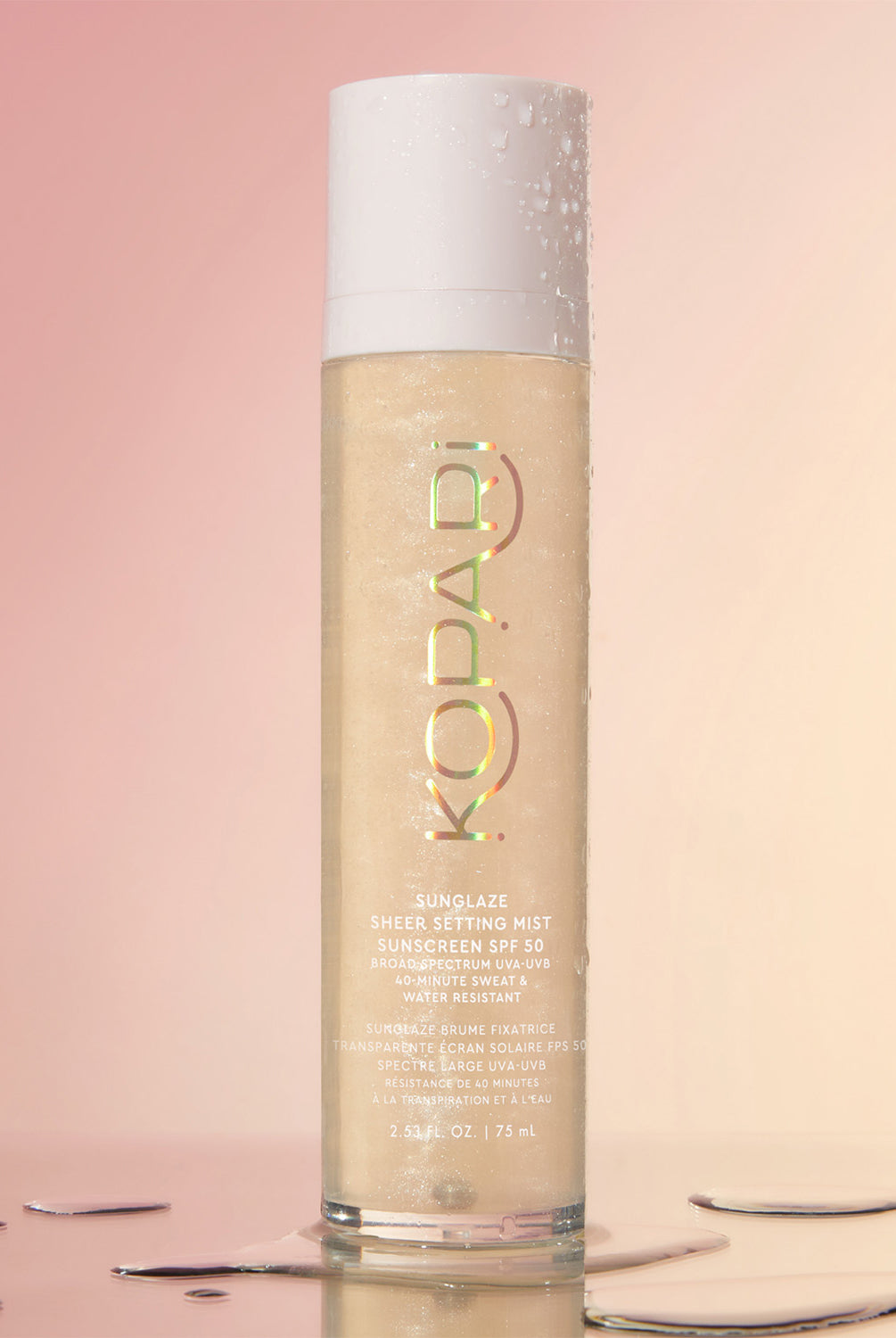 A close-up of the Kopari Sun Shield Body Glow SPF 50 spray bottle standing upright with water droplets on a pink background.