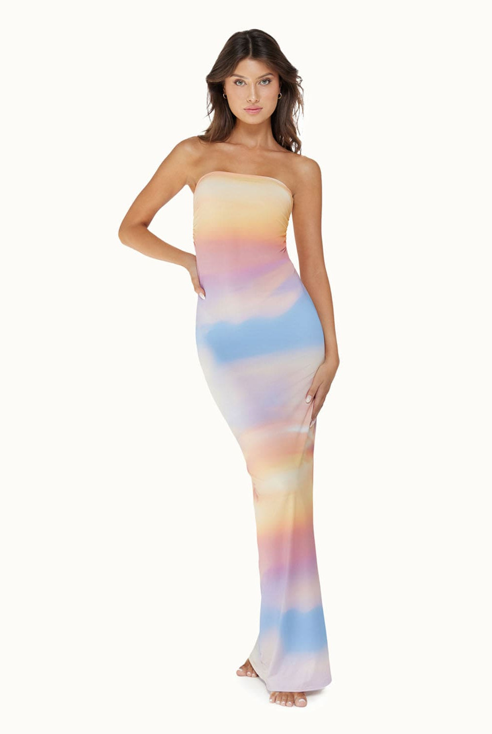 A brunette woman wearing a multi colored floor length dress standing in front of a white wall. 