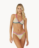 A blonde woman wearing a tan bikini stands in front of a white wall.