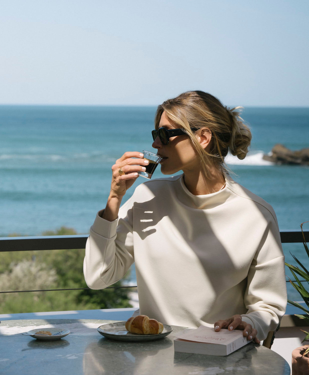 Woman drinking coffee by the ocean wearing a beige top