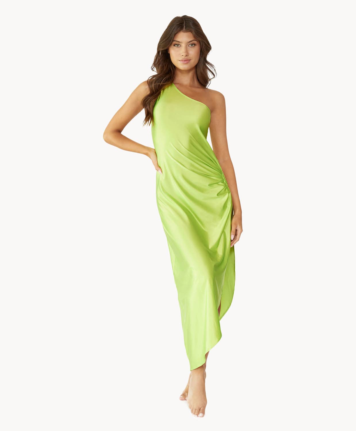 Brunette woman wearing a lime green one-shoulder dress with side slit and ring detail stands in front of white wall.