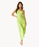 Brunette woman wearing a lime green one-shoulder dress with side slit and ring detail stands in front of white wall.