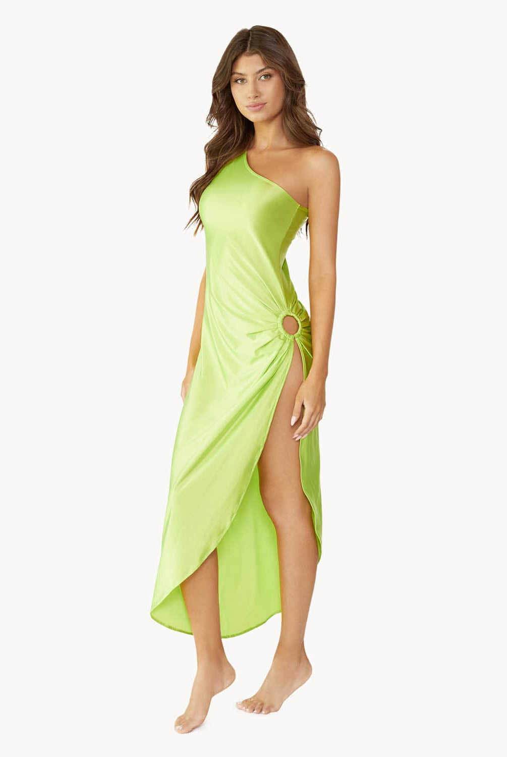 Brunette woman wearing a lime green one-shoulder dress with side slit and ring detail stands in front of white wall.