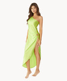 Brunette woman wearing a lime green one-shoulder dress with side slit and ring detail stands in front of white wall.