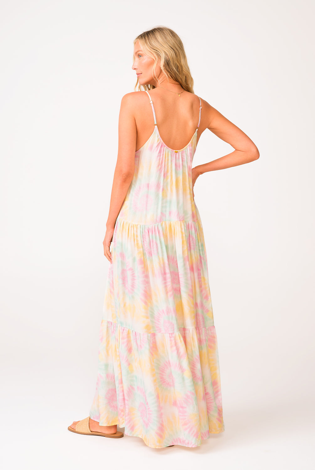 A woman wearing a tie dye long dress facing towards a white wall.