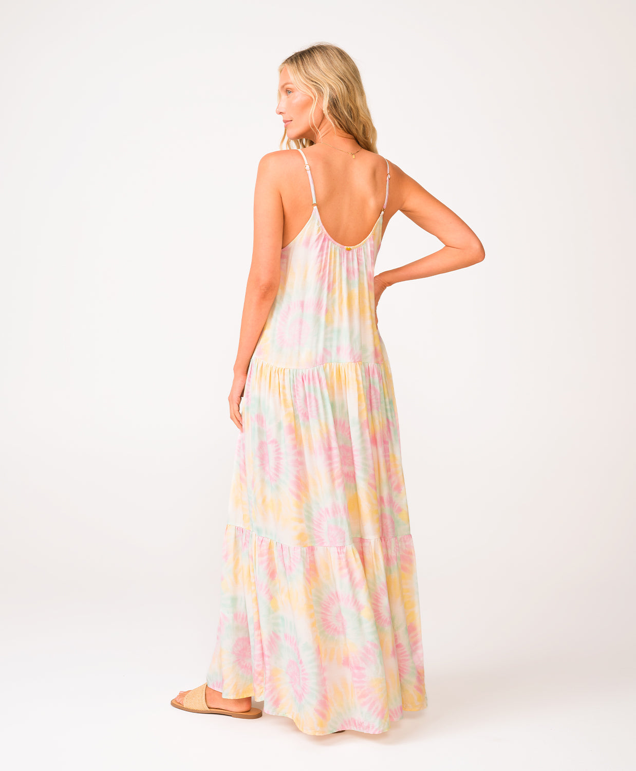 A woman wearing a tie dye long dress facing towards a white wall.