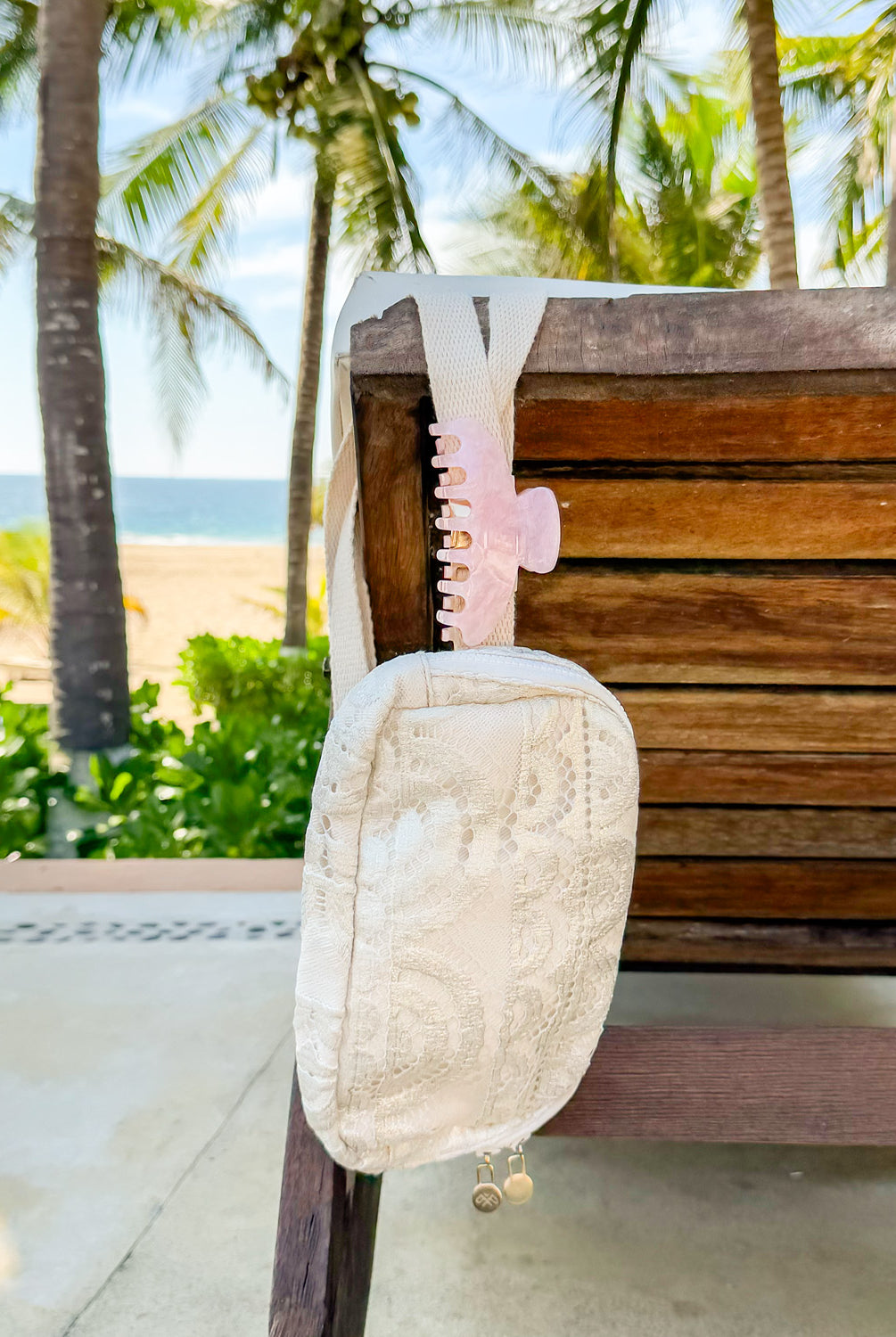 A cream colored lace crossbody purse and a pink hair clip hanging on a bench outside.