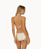 A blonde woman wearing an ivory lace bikini facing towards a white wall.