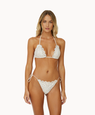 A blonde woman wearing an ivory lace bikini standing against a white wall.
